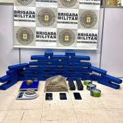Tabletes de maconha, diversas porções de drogas, uma balança de precisão, três aparelhos celulares, banner da BM ao fundo 