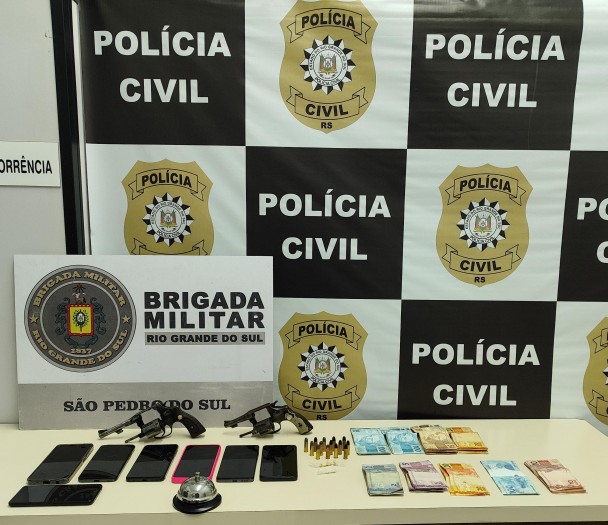 materiais apreendidos, armas, munições, dinheiro, drogas, celulares, banner da BM e da PC ao fundo 