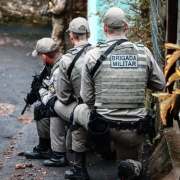 três homens PMs agachados e armados e mais a frente um PM também armado 