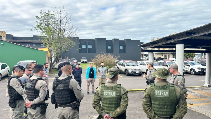 policiais militares em pé, numa roda, momento de instruções da operçaão. em um estacionamento, céu com  nuvens. 