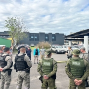 policiais militares em pé, numa roda, momento de instruções da operçaão. em um estacionamento, céu com  nuvens. 