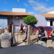 quatro policiais, sendo três militares e um civil, em frente a uma moradia humilde, tem pessoas na área da casa, sol e céu azul 