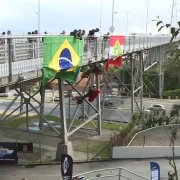 ponte com bandeiras do Brasil e de SC e vê-se o PM carregando a vítima nas costas durante a prova, as cordas içando os PMs e outros sobre a ponte