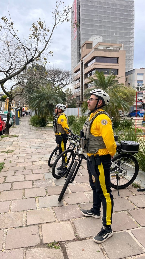 dois policiais militares que patrulham com bicicletas em prontidão numa calçada arborizada e prédios aparecem ao fundo