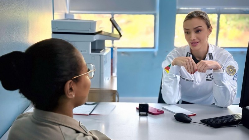 consulta no hospital da BM, uma PM feminina está em frente a médica entrevistada, que está atrás da mesa. ambas estão sentadas e vestindo as fardas, a da médica é branca.