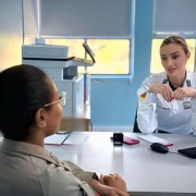 consulta no hospital da BM, uma PM feminina está em frente a médica entrevistada, que está atrás da mesa. ambas estão sentadas e vestindo as fardas, a da médica é branca. 