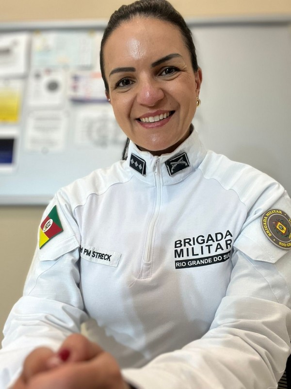 PM está sentada, com a frada branca, cabelo preso, sorrindo, mãos à frente, ao fundo aparece um quadro branco com avisos
