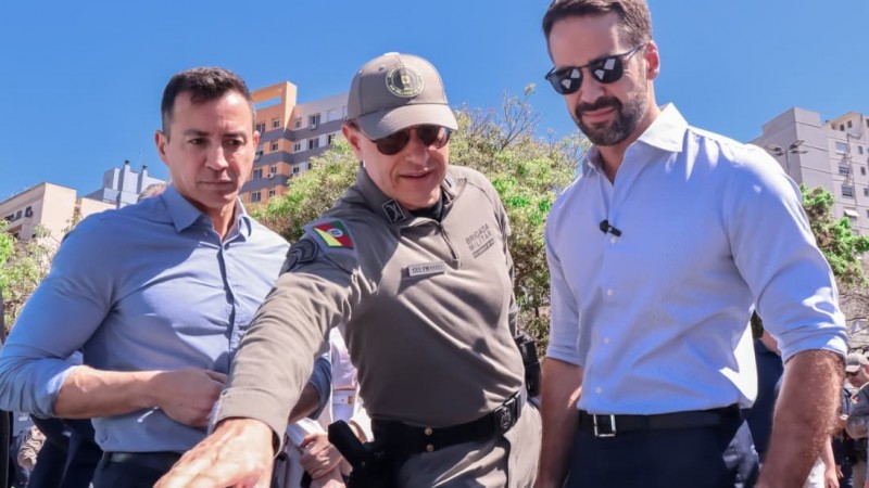 As três autoridades aparecem de frente conferindo as novas armas, ao fundo aparece o céu azul e prédios do Centro Histórico