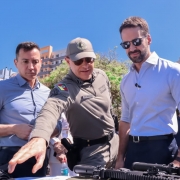 As três autoridades aparecem de frente conferindo as novas armas, ao fundo aparece o céu azul e prédios do Centro Histórico