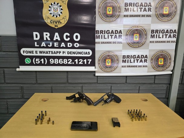 Armas e municções aparecem sobre uma mesa. Ao fundo, estão dois banners, sendo um da DRACO e outro da BM.