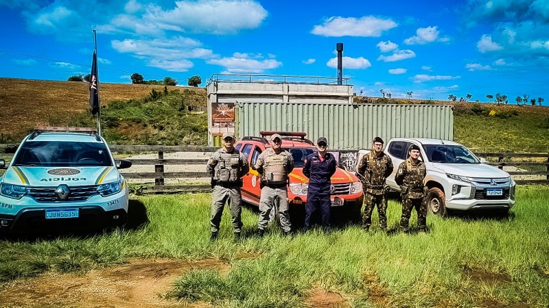 Militares aparecem em uma área de campo aberto e verde, em um dia ensolarado, céu azul. Atrás deles, aparecem as viaturas, sendo da BM, dos Bombeiros e do Exército. Aparece ao fundo um conteiner e uma paisagem aberta.