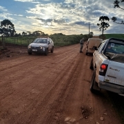 Três veículos aparecem em uma estrada de chão, à frente um PM. 