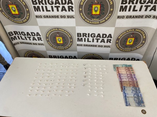 Porções de drogas e cédulas de dinheiro distribuídos sobre uma mesa. Ao fundo, um banner com os brasões da BM
