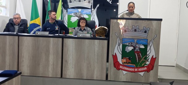 PM homenageada falando no púlpito da Câmara, ao lado de vereadores sentados atrás à Mesa Diretora