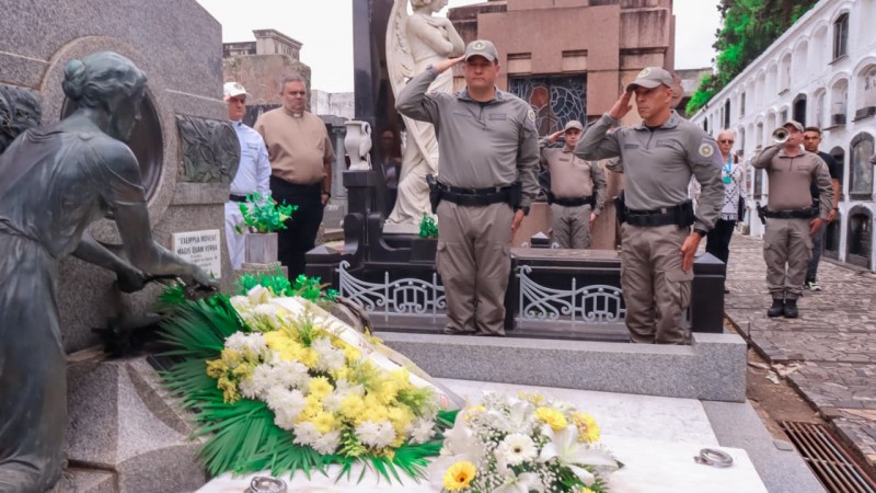 Militares e civis estão em torno do jazigo ornamentado com flores. Aparece dois PMs, Chefe do EMBM e o Subcomandante-Geral da BM prestando continência ao homenageado