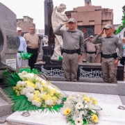 Militares e civis estão em torno do jazigo ornamentado com flores. Aparece dois PMs, Chefe do EMBM e o Subcomandante-Geral da BM prestando continência ao homenageado