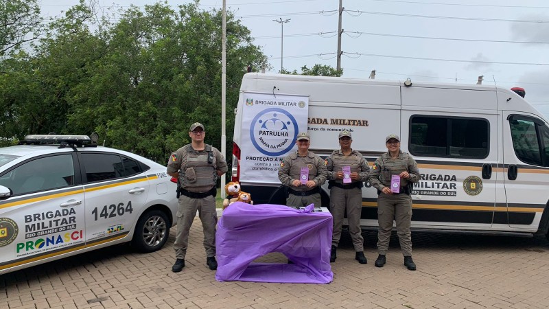 PMs aparecem em pé em frente a duas viaturas, sendo um automóvel e um micro-ônibus, segurando folders da Patrulha Maria da Penha