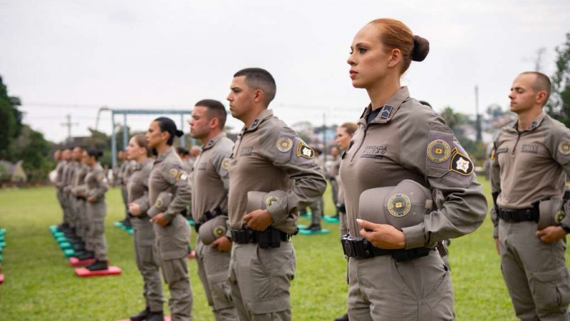PM feminina em primeiro plano. Fileiras de PMs dispostos no campo da APM durante solenidade