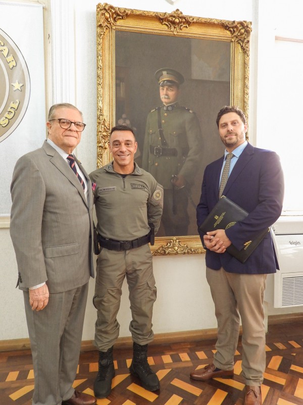 Os três homens estão em pé e em frente ao quadro de parede com a imagem do Patrono da BM