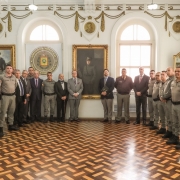 Militares e membros do Conselho Superior da BM formam um semicírculo. No centro está o quadro com a imagem do Coronel Massot e o neto e o bisneto, um de cada lado do Patrono da Instituição.  