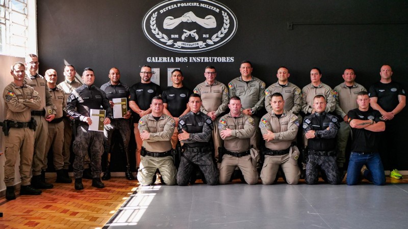 PMs aparecem em duas fileiras, fardados. Dois deles seguram o Manual impresso. Ao fundo, lê-se na parede preta: Galeria dos Instrutores. Há também o brevê da Defesa Policial Militar.