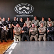 PMs aparecem em duas fileiras, fardados. Dois deles seguram o Manual impresso. Ao fundo, lê-se na parede preta: Galeria dos Instrutores. Há também o brevê da Defesa Policial Militar.  