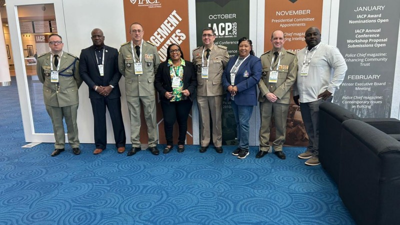 Foto mostra os quatro integrantes da comitiva da Brigada Militar posando para foto junto a uma comitiva estrangeira. Por trás deles, estão cartazes da feira IACP 2025