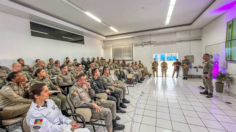 Uma sala de aula está repleta de formandos do Curso. À frente e em pé, fala o diretor do Departamento de Ensino. Há luzes cor-de-rosa decorando o ambiente em alusão ao tema da proteção às mulheres. 