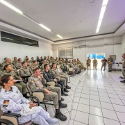 Uma sala de aula está repleta de formandos do Curso. À frente e em pé, fala o diretor do Departamento de Ensino. Há luzes cor-de-rosa decorando o ambiente em alusão ao tema da proteção às mulheres. 