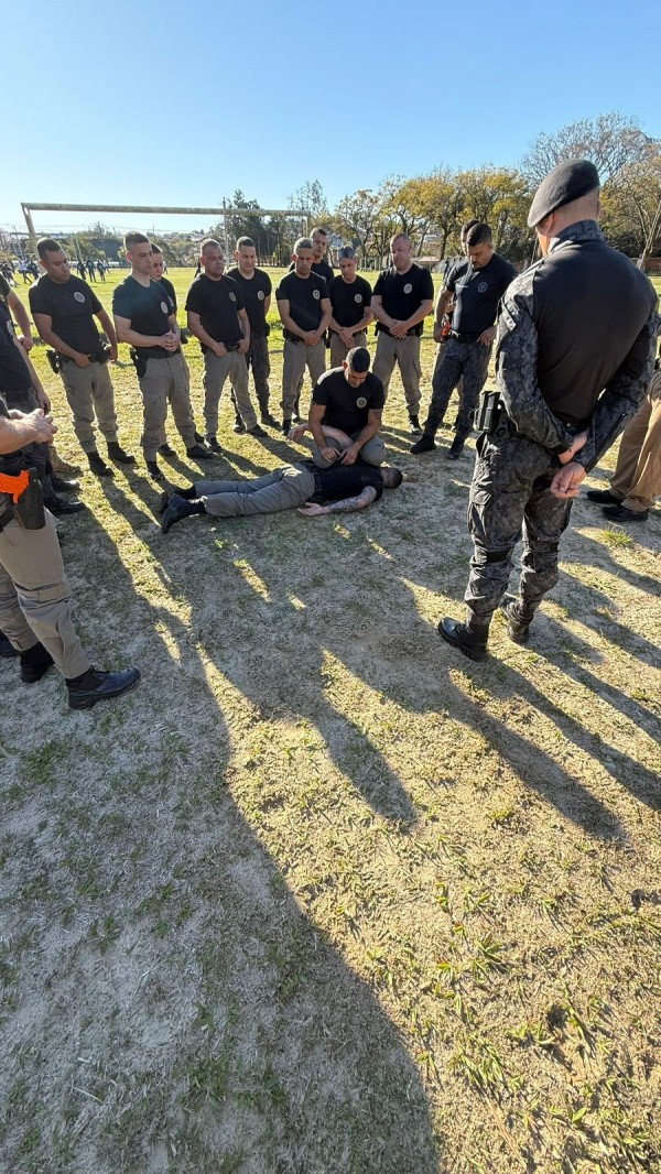 PMs em pé e em círculo observando uma técnica de algemação entre dois PMs, sendo um deitado de bruços e outro ajoelhado.
