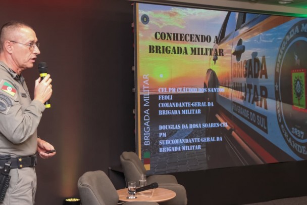 Comandante-Geral aparece de lado, com o microfone em mãos e diante de um slide que traz as informações sobre a BM. 