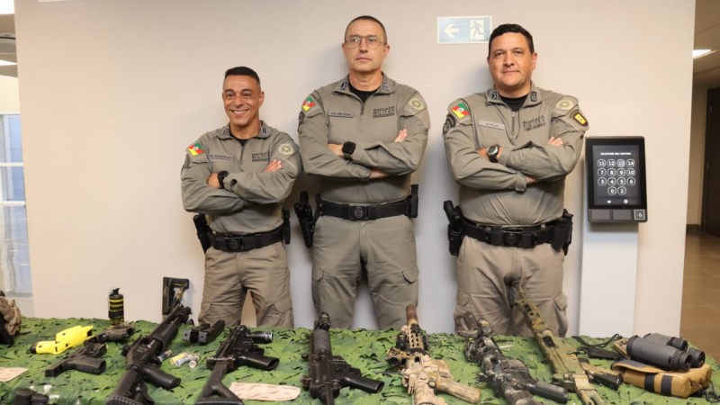 Os três comandantes da Brigada Militar aparecem em pé, com os braços cruzados, em frente a uma mesa com armas expostas e uma parede clara ao fundo. 