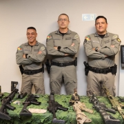 Os três comandantes da Brigada Militar aparecem em pé, com os braços cruzados, em frente a uma mesa com armas expostas e uma parede clara ao fundo. 