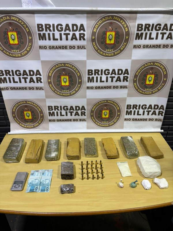 Tabletes de maconha e outros materiais aparecem sobre uma mesa e ao fundo está o banner com o brasão da BM. 