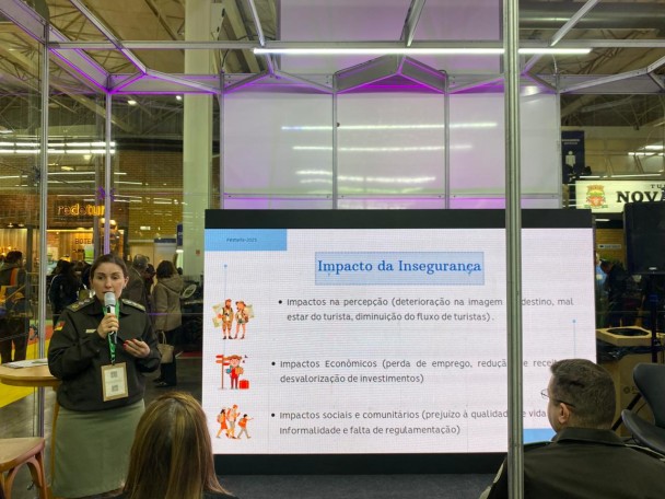 PM feminina aparece falando ao microfone durante a palestra que ministrou no evento. Há um slide que aborda o tema do "Impacto da Insegurança" e público no ginásio bem como assistindo a palestra. 