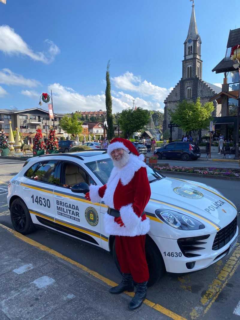 Papai Noel aparece em frente a uma viatura. Ao fundo vê-se a decoração natalina das ruas e uma igreja.  