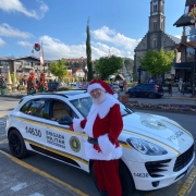 Papai Noel aparece em frente a uma viatura. Ao fundo vê-se a decoração natalina das ruas e uma igreja.  
