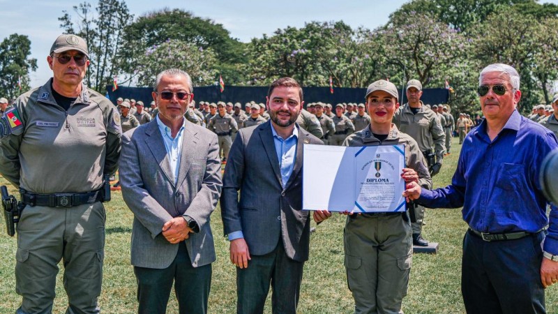 No campo do estádio, aparecem da esquerda para a direita, o Comandante-Geral, o Secretário de Segurança Pública, o Vice-Governador, a Soldado PM condecorada e seu padrinho. 
