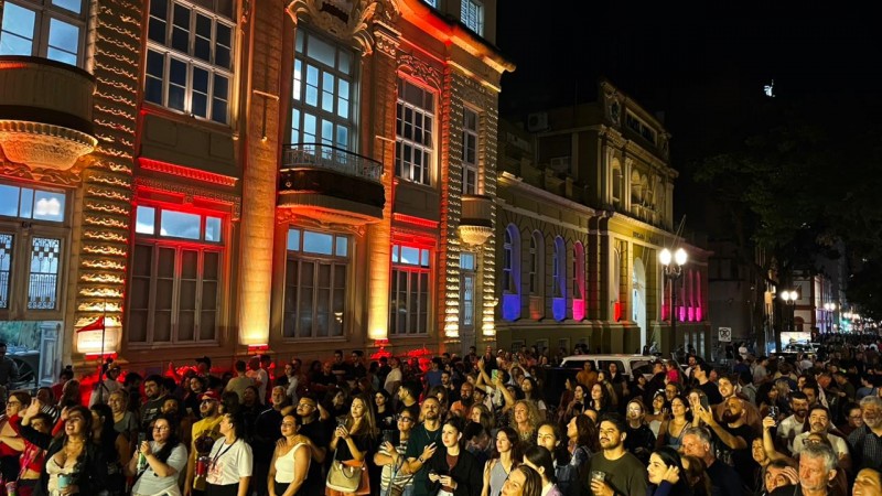 O público ocupa a rua em frente ao Museu da Brigada Militar enquanto assiste uma  apresentação musical. Os prédios do Museu e do Comando-geral estão iluminados com corrs variadas. 