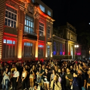 O público ocupa a rua em frente ao Museu da Brigada Militar enquanto assiste uma  apresentação musical. Os prédios do Museu e do Comando-geral estão iluminados com corrs variadas. 