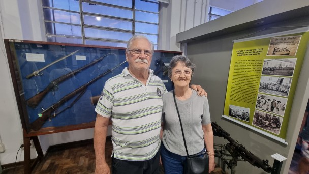 O casal aparece abraçado em frente a um painel com armas armas antigas. 
