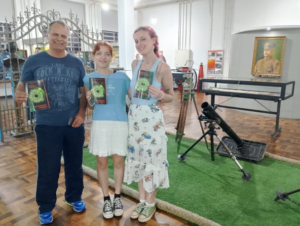 As três pessoas aparecem segurando o mapa do evento Noite dos Museus e, ao fundo, aparecem objetos da sala de exposição do segundo andar do Museu da Brigada. 