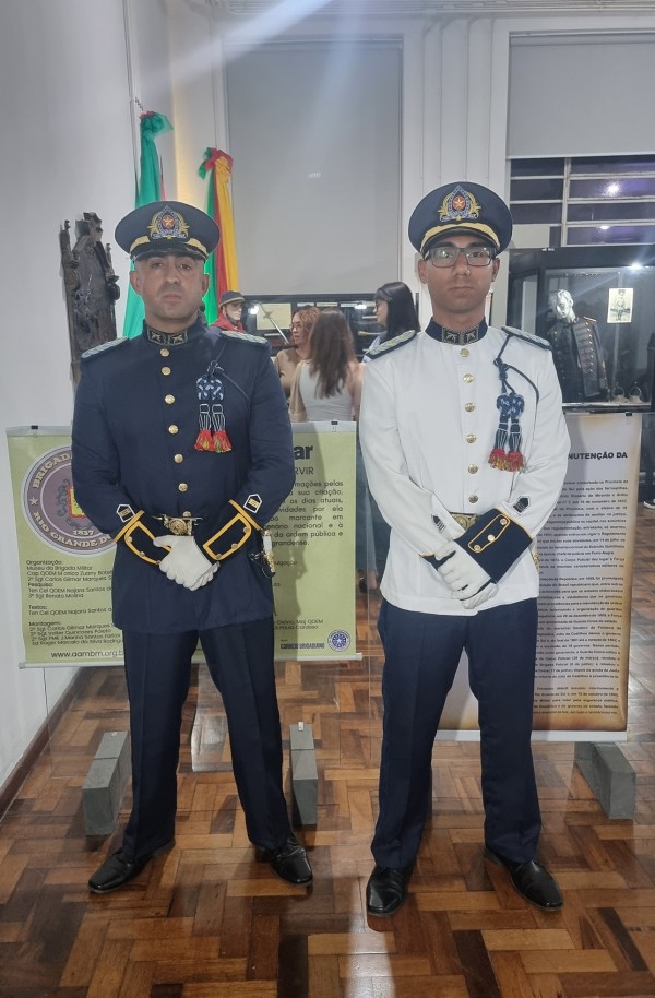 A dupla de PMs aparece trajando as duas variações da tradicional farda, sendo a primeira toda azul e a segunda com a túnica branca. Ao fundo, vê-se objetos do Museu e visitantes. 