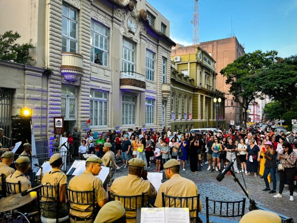 Os músicos da Banda estão sobre o palco montado em frente ao Museu. O público acompanha a apresentação em frente, na rua dos Andradas, endereço do Museu e da sede do Comando-Geral. 