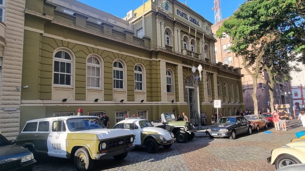 Diante da fachada da sede do Comando-Geral aparecem os carros estacionados que foram utilizados pelo efetivo no século passado. 