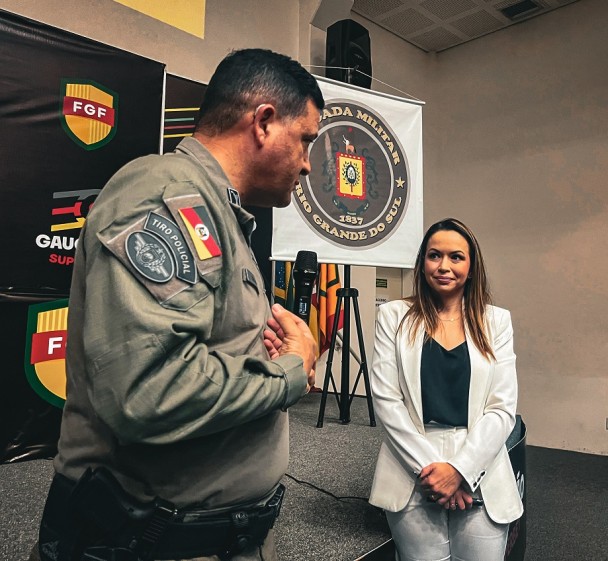 A dupla aparece diante do palco, com o painel da Federação Gaúcha de Futebol ao fundo assim como o banner da BM. 