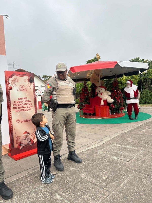 O PM aparece conversando com uma criança de cerca de 5 anos. Ao fundo, aparecem os adereços natalinos, como o Papai Noel. 