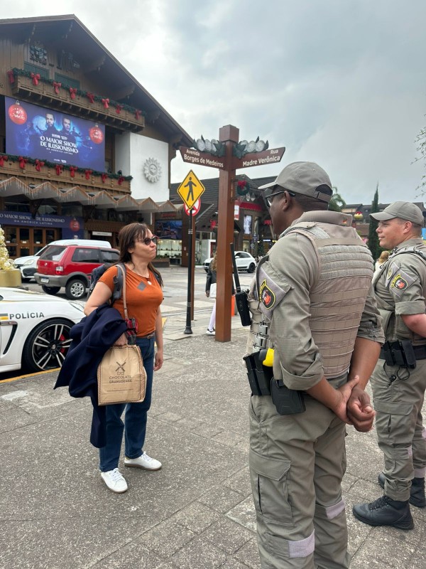 PMs aparecem conversando com uma senhora, que segura uma sacola e casaco. O céu está nublado e ao fundo aparece um ginásio de exposições.  