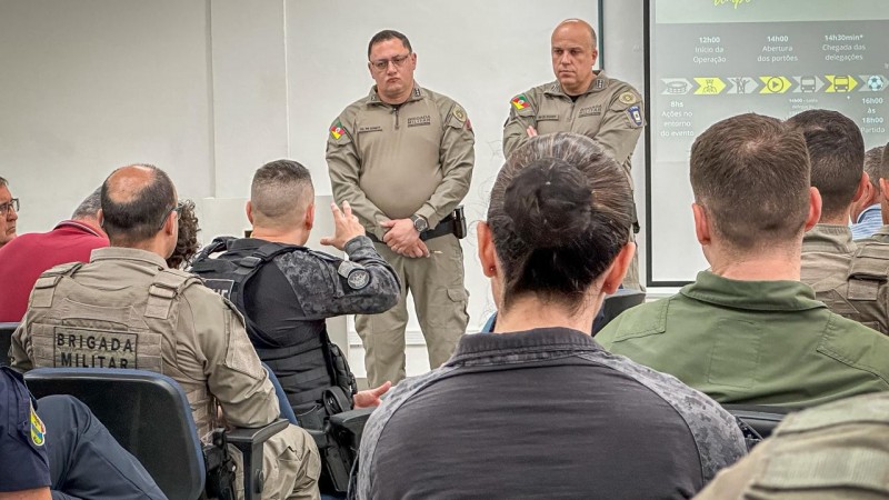 Dupla de PMs aparece em pé diante de um público de PMs ouvindo a apresentação do cenário operacional. 