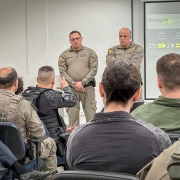 Dupla de PMs aparece em pé diante de um público de PMs ouvindo a apresentação do cenário operacional. 